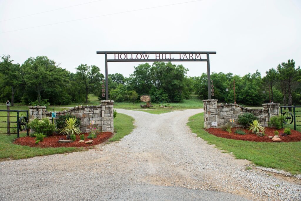 Hollow Hill Event Center Wedding and Event Venue, Weatherford, Texas. Wooded area with iron sculpture and fire pit in sunken sand area. Metal sign with venue name stretched over gravel road between 2 stone walls. Trees and field in background. Red mulched landscaped beds in front of stone walls.