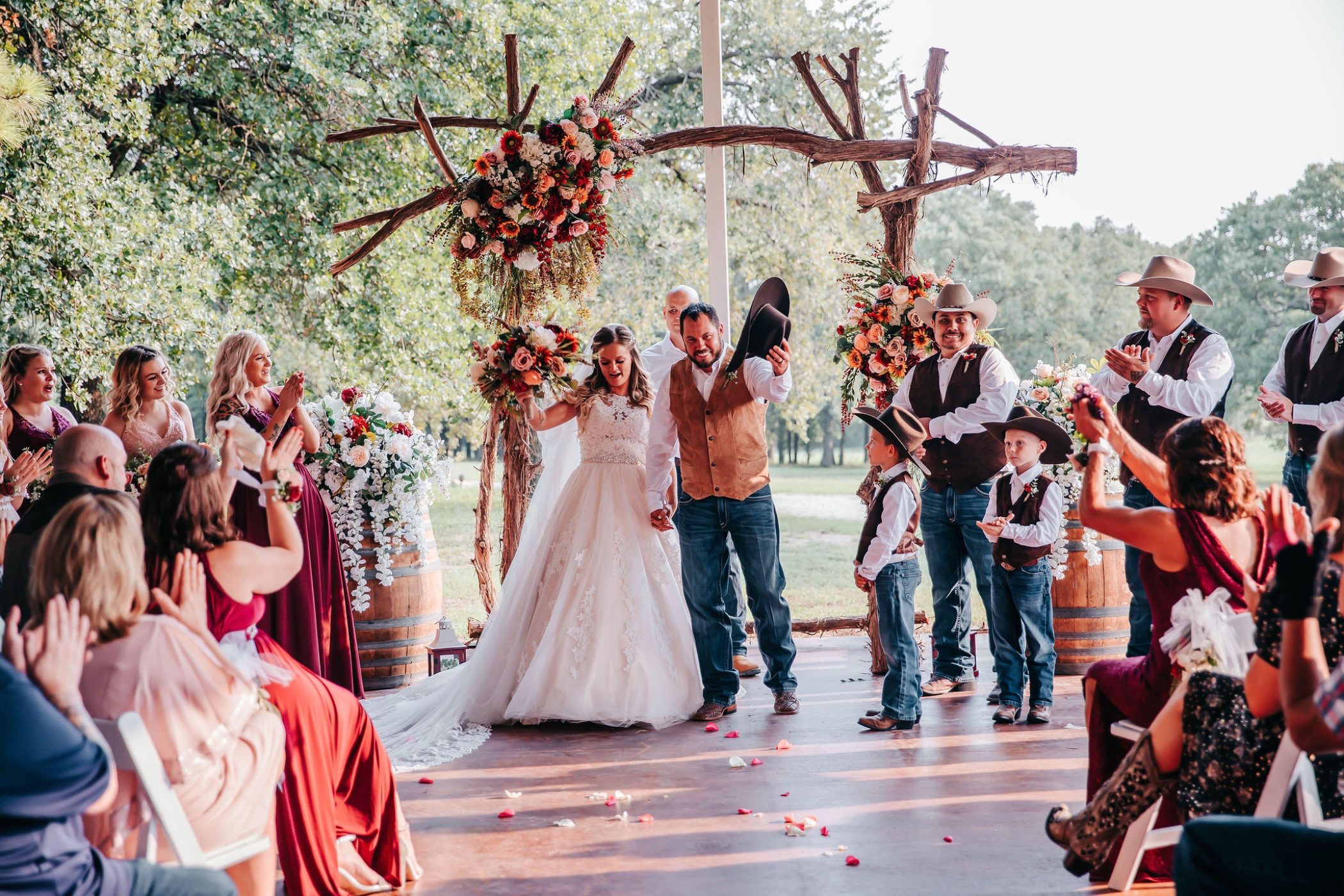 Hollow Hill Event Center Wedding and Event Venue, Weatherford, Texas. Bride and groom celebrating end of ceremony under log arch with floral decor.