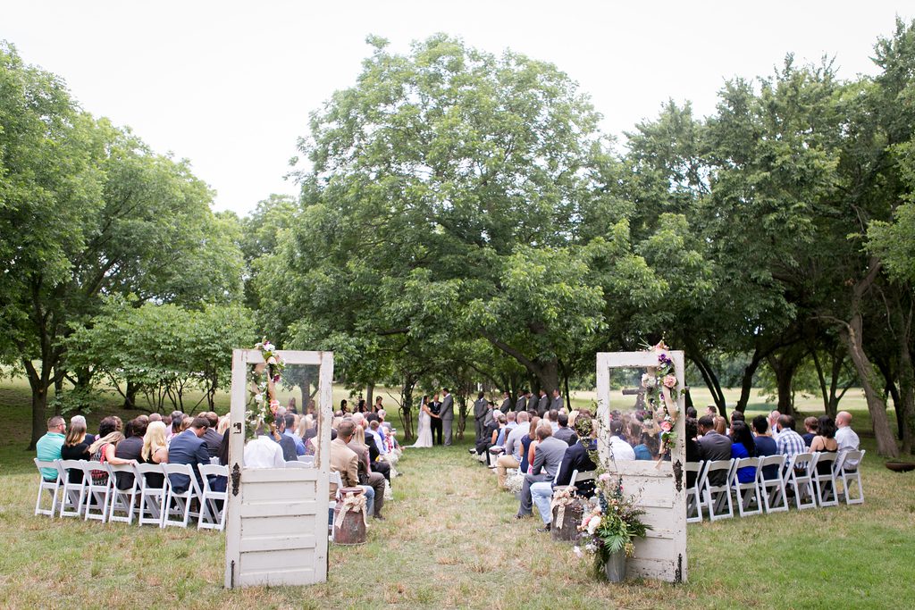 Hollow Hill Event Center Wedding and Event Venue, Weatherford, Texas. Wedding ceremony in field surrounded by trees. Old screen doors with floral arrangements on them at entrance.