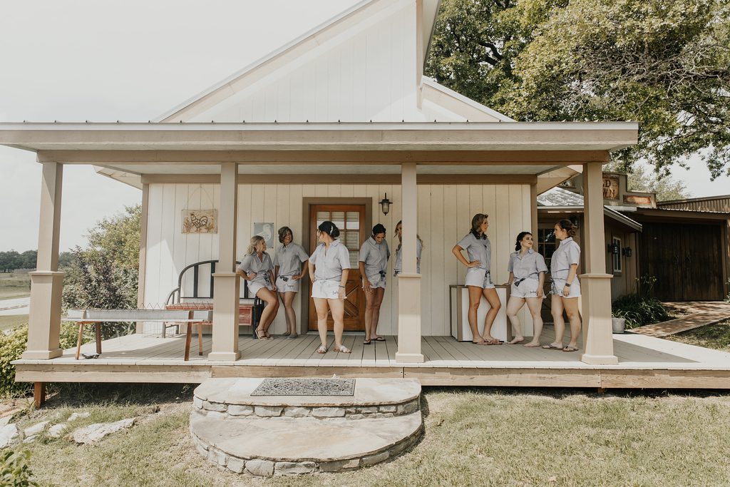 Hollow Hill Event Center Wedding and Event Venue, Weatherford, Texas. Bridal party standing on porch of cream colored building smiling and laughing.